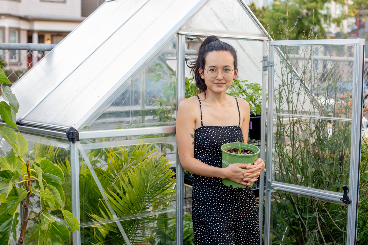 Melati Citrawireja standing in front of a small greenhouse holding a potted plant and looking at the camera