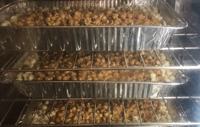 Three trays of caramel coated popcorn in a commercial oven, viewed through the glass of the oven door.