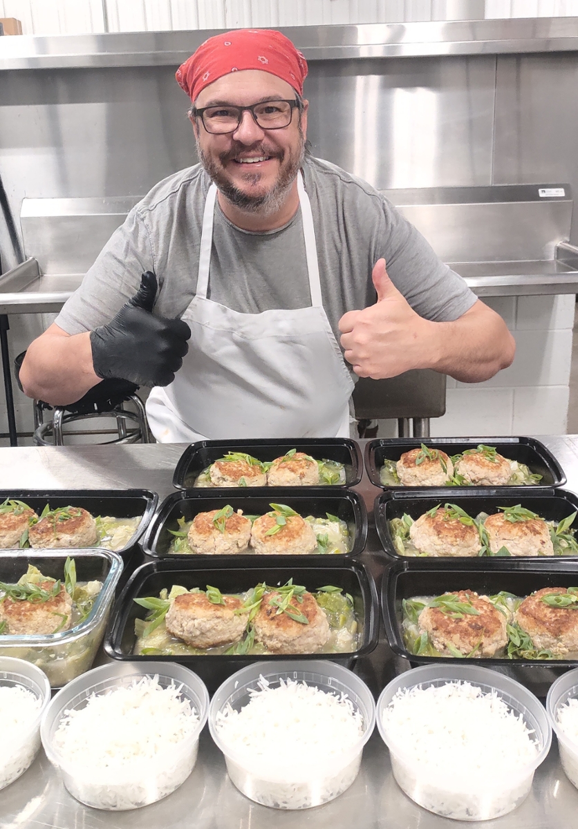 A man wearing a red bandana and white apron gives a thumbs-up in front of several prepared and packaged meals of meatballs over vegetables 