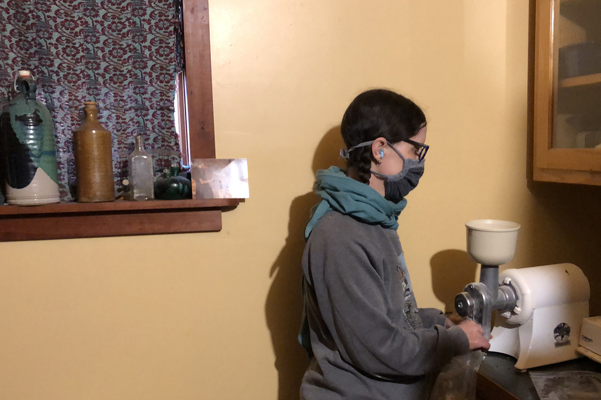 Denise Breeden Ost in profile with face mask holding a back under a grain grinder attached to a motorized juicer in a room with woodwork and a yellow wall.