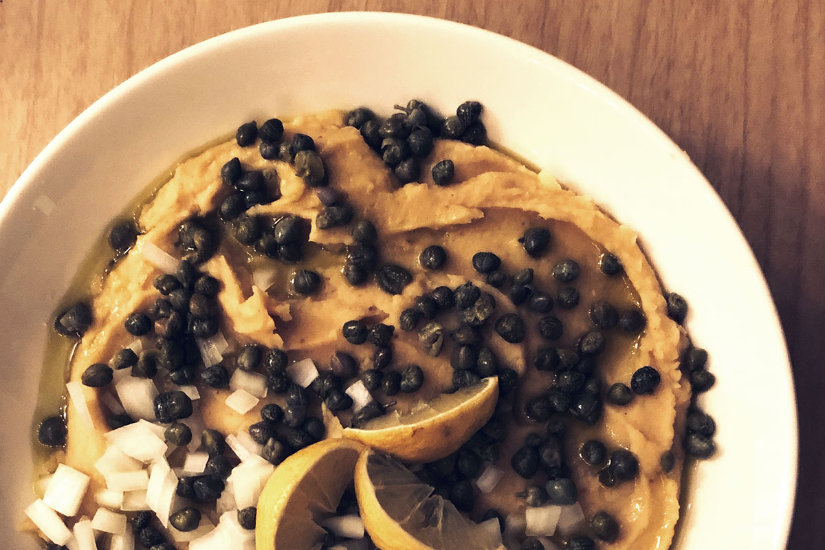 close up of a beige-colored dip in a bowl with capers scattered over it and lemon wedges