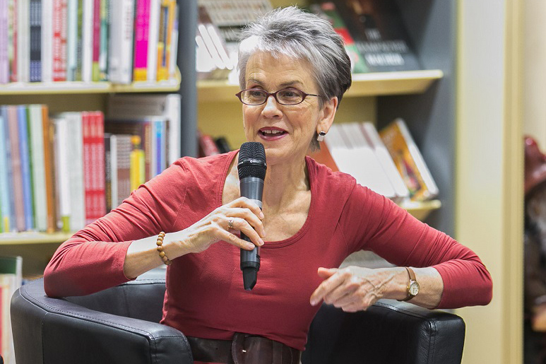 Frances Moore Lappé sitting in an armchair holding a microphone and smiling, arms raised as if in animated speech. Bookshelves in the background of a well lit room