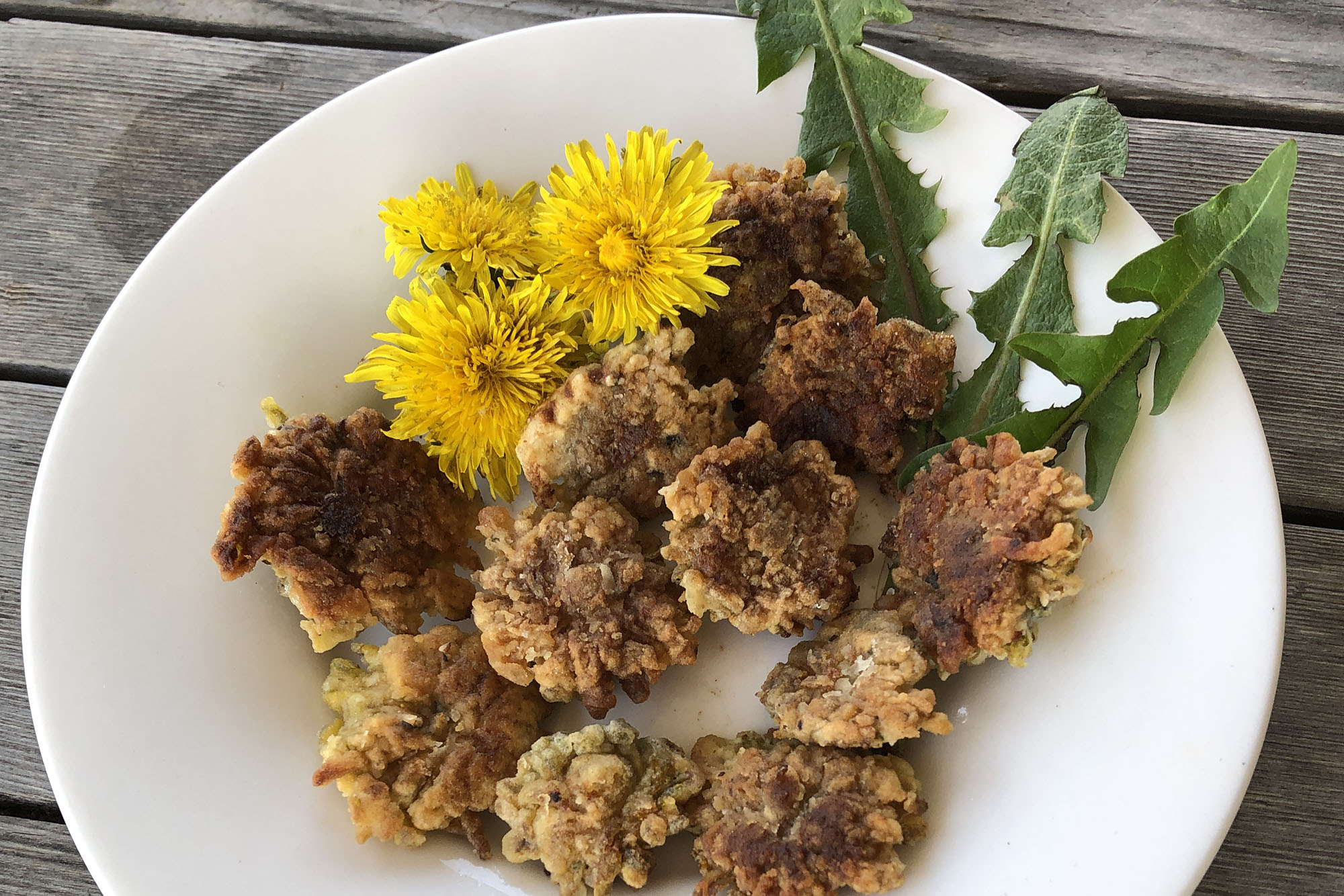 round fried foods on a white plate with yellow dandelion flowers and few jagged leaves