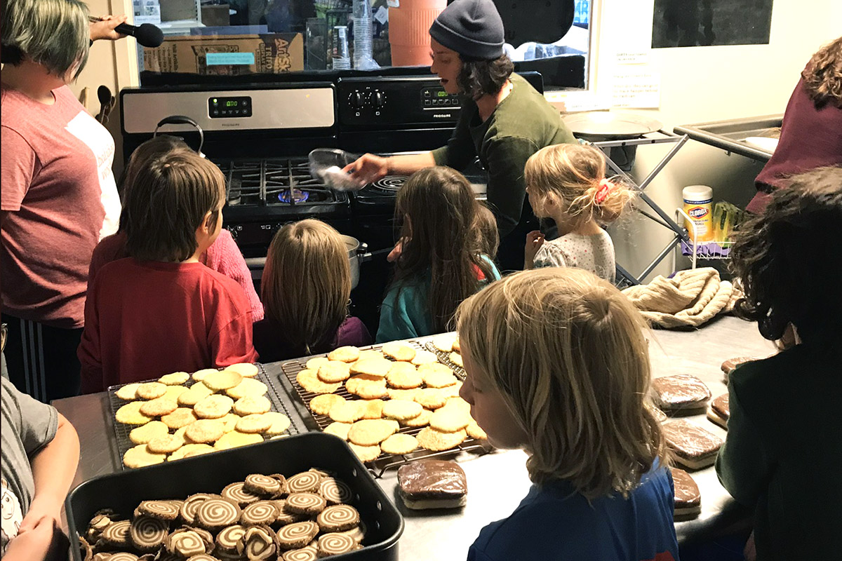 A group of children gathered around a stove with a woman showing them a bowl. Cookies in the foreground
