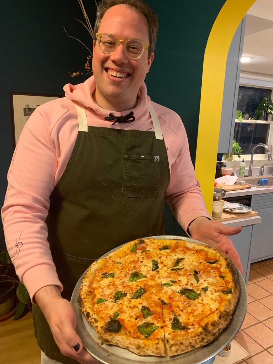 Pete Giordano holding a pizza and smiling at the camera with a domestic setting in the background