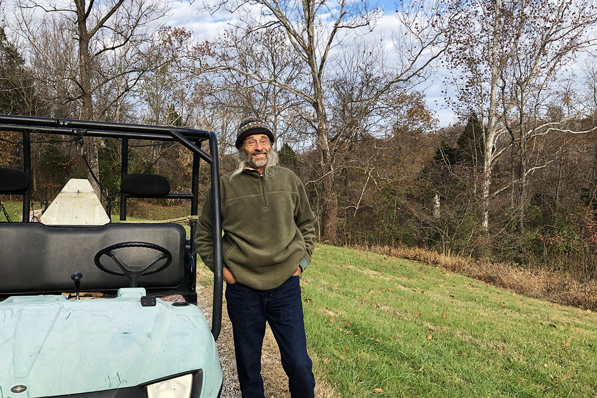Larry Gillen standing next to a faded blue and black jeep-like vehicle with bare trees and brush in the background