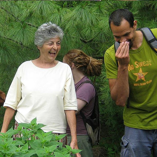 Lucille Bertuccio laughing, Ross Gay sniffing a plant, outside in front of evergreen trees