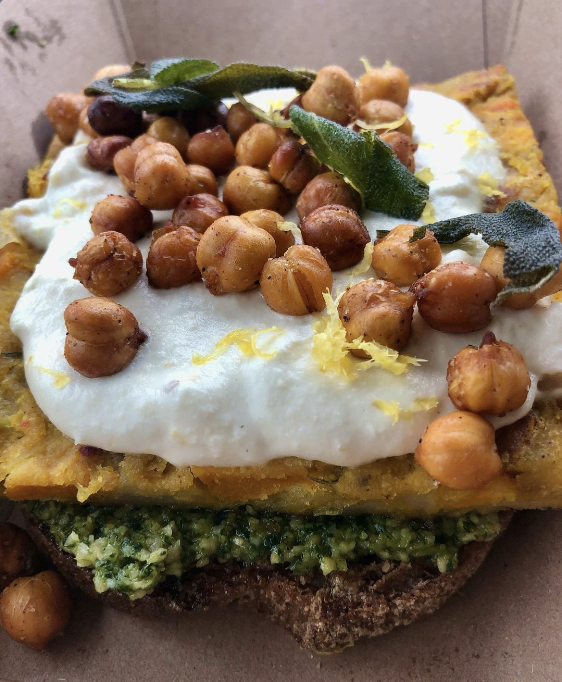 roasted chickpeas on top of a white, creamy layer, on top of a layer of somethng yellow and sliced on top of a layer of pesto on top of a rustic bread. The dish is garnished with lemon zest and sage leaves. 