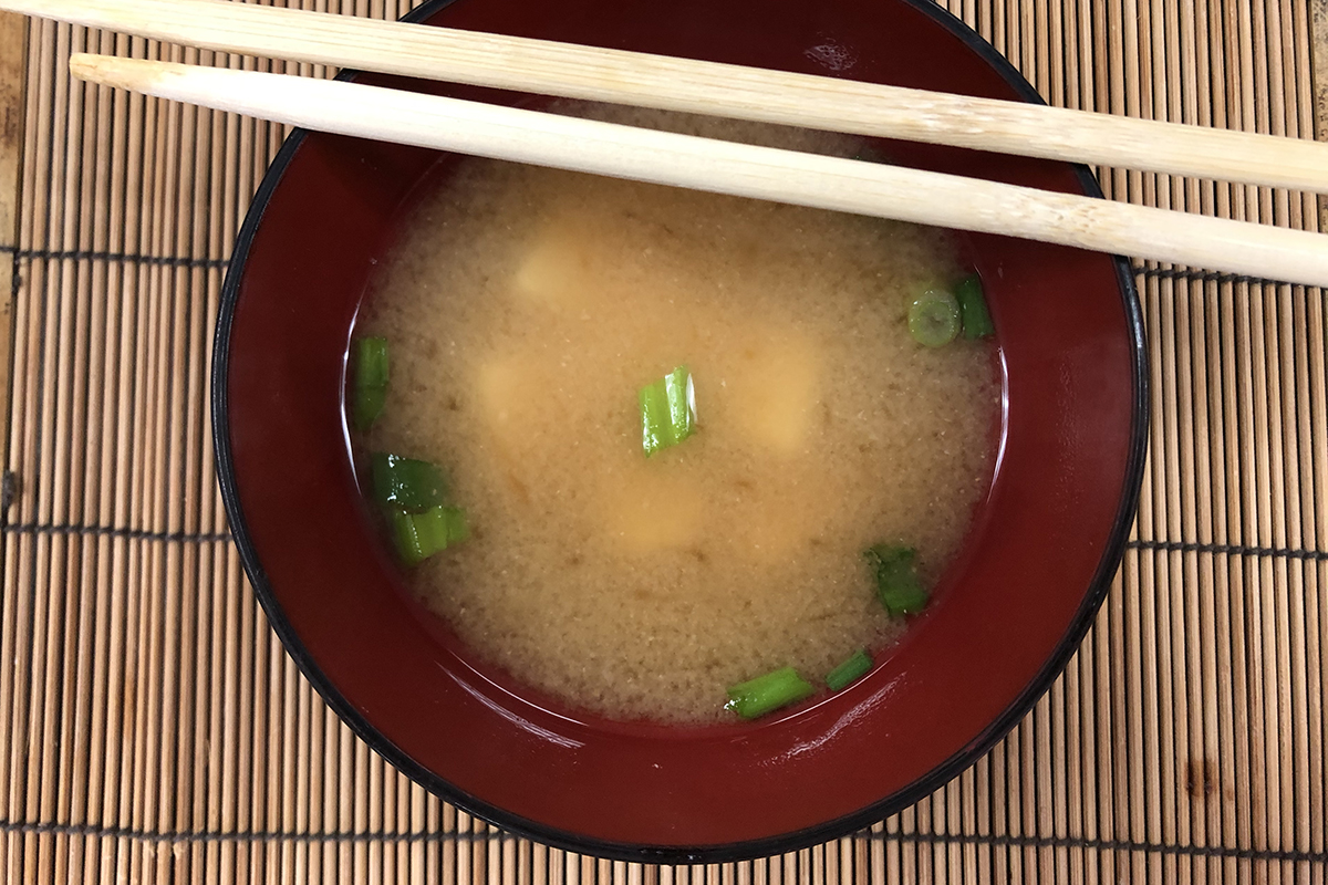 A bowl with cloudy broth and green bits, overhead view, bowl has red interior and two pale sticks are across the bowl towards the top. 