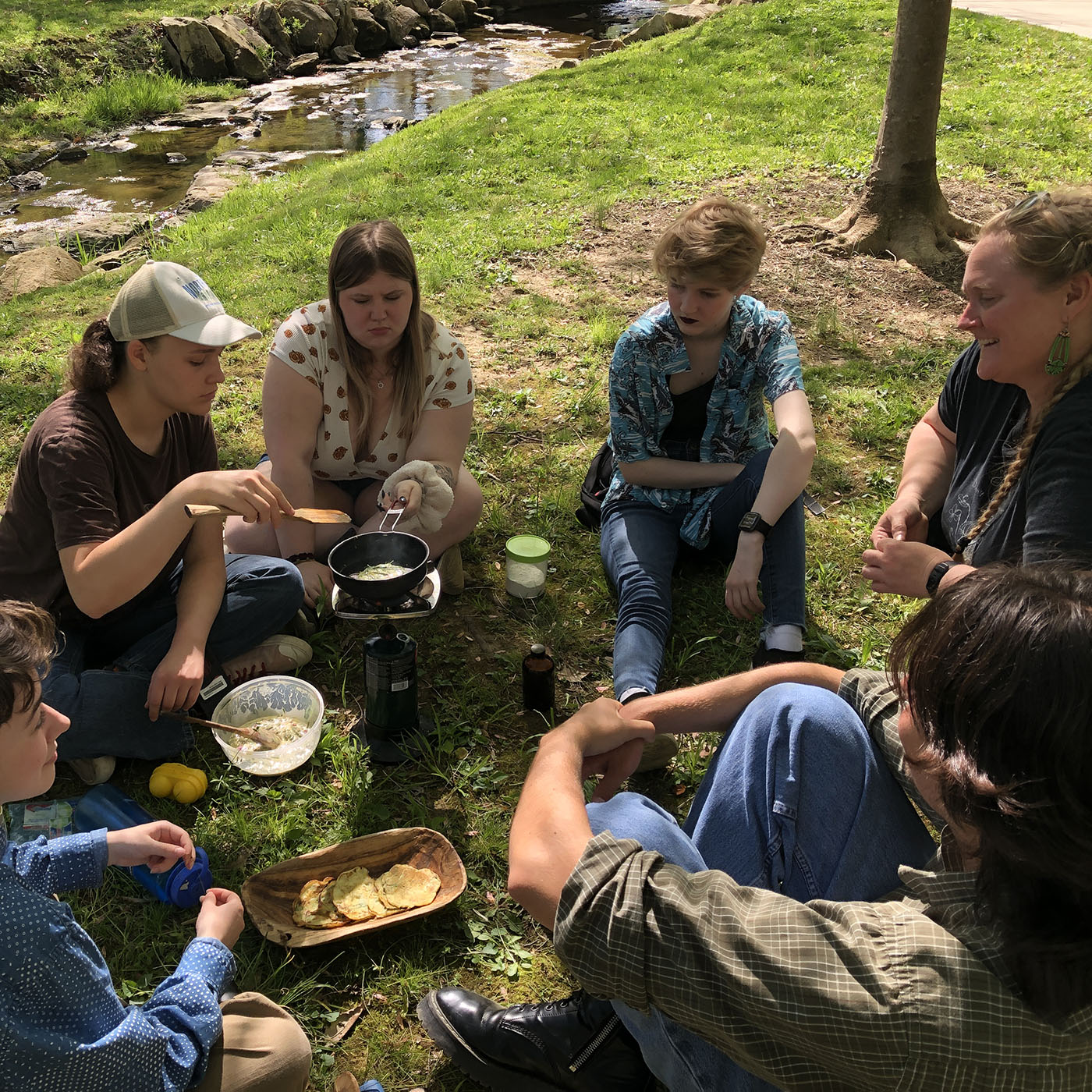 Several young people sitting around a campstove with a skilled, next to a creek. A wooden bowl with pancakes is visible, and other ingredients.