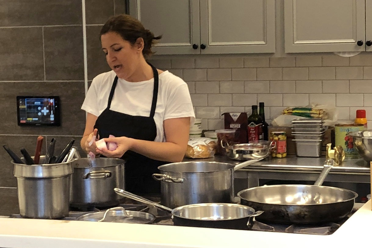 Nafsika Papacharalampous standing in comercial kitchen with pots and pans, cutting food into a pot