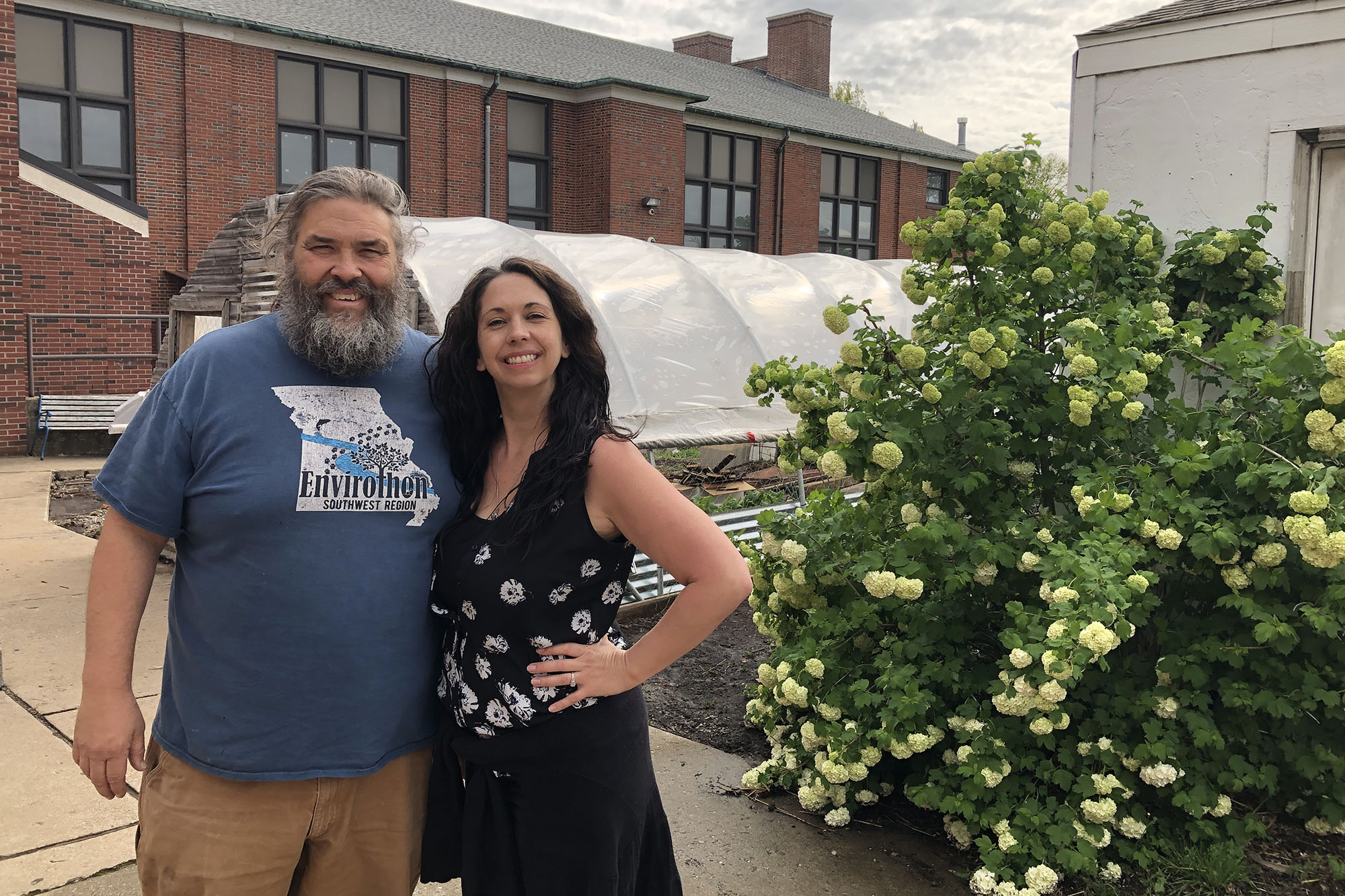 Paul Eps and Brandi Nelson standing in front of a high tunnel and a brick building with a flowering bush next to them. They are smiling at the camera