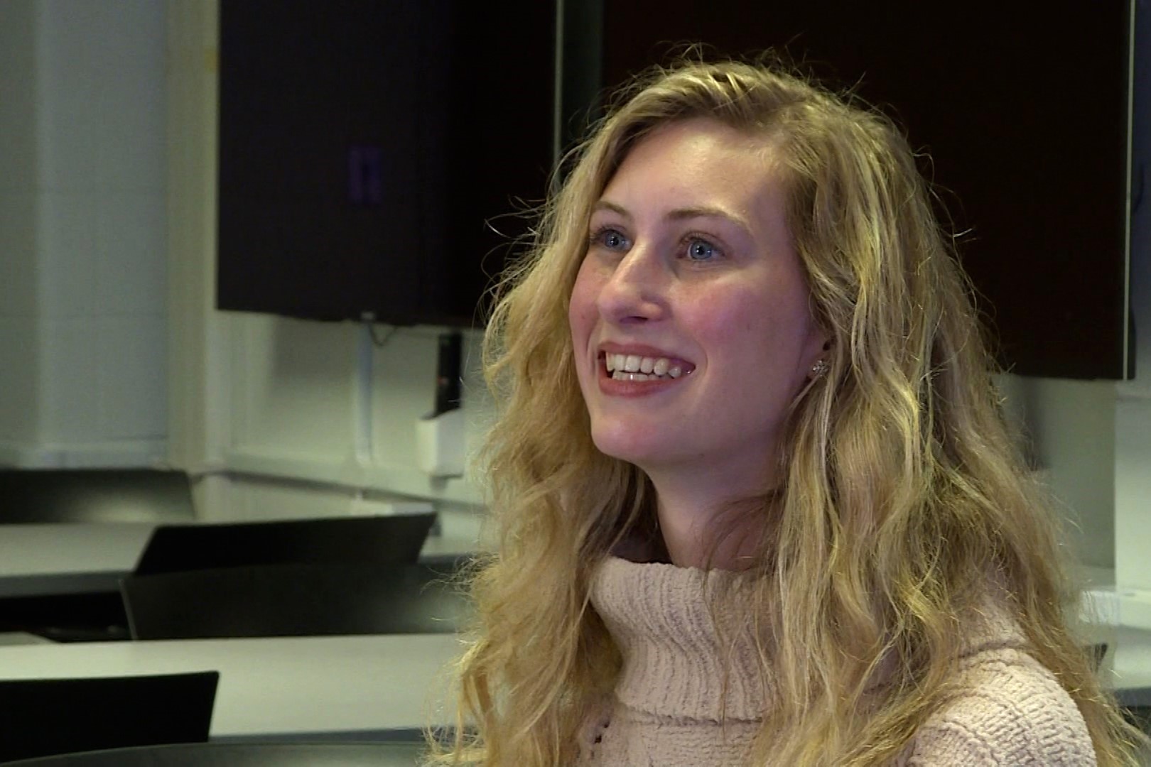 Paige Moore, wearing a light pink turtle neck sweater, smiles at her ASL interpreter.