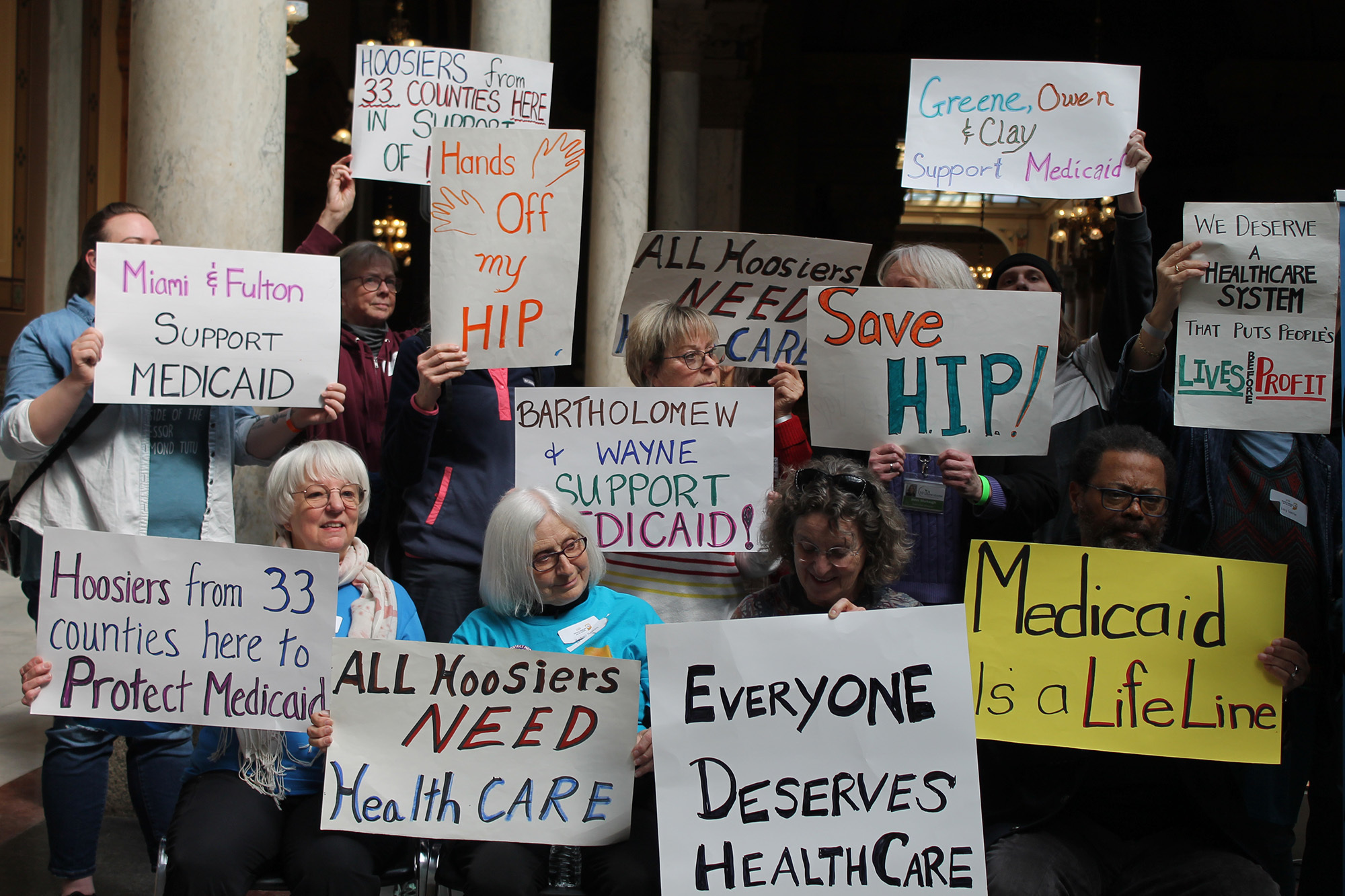 People gathered at a rally inside the Statehouse holding signs that say “Hoosiers from 33 counties here to Protect Medicaid,” “Hands off my HIP,” “All Hoosiers need Healthcare,” “Everyone deserves healthcare,” “Medicaid is a lifeline,” “Save HIP,” “W