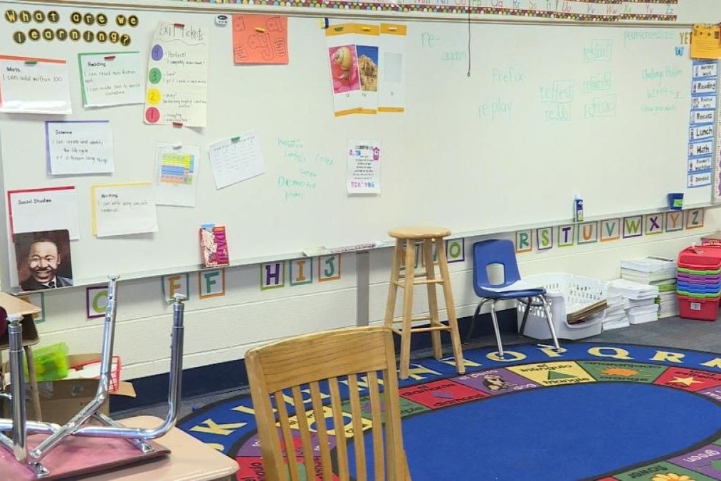 Empty elementary school classroom with a colorful round rug and a whiteboard with writing and posters.