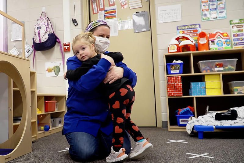 A child receives a hug from a child care worker wearing a mask.
