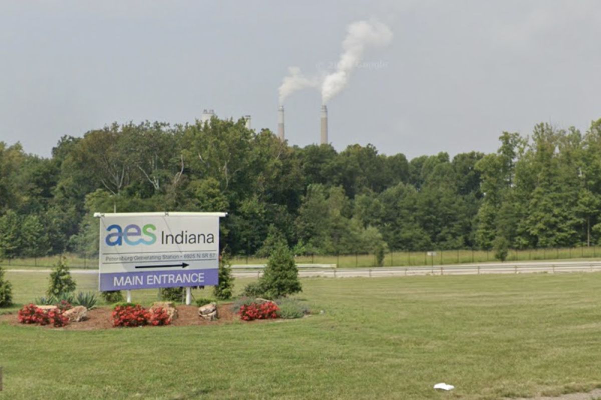 The entrance sign to AES Indiana's Petersburg Generating Station. In the background of the image are a line of trees and two smoke stacks stretching into the sky behind them.