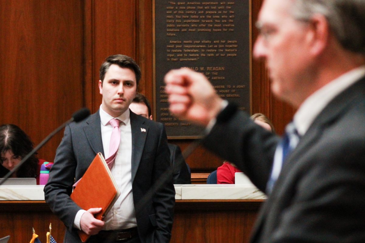 Andrew Ireland stands on the House floor looking at Matt Pierce, who is in the foreground, standing in profile and raising his hand while he speaks. Ireland is a White man with dark hair, wearing a suit and tie. Pierce is a White man with gray hair