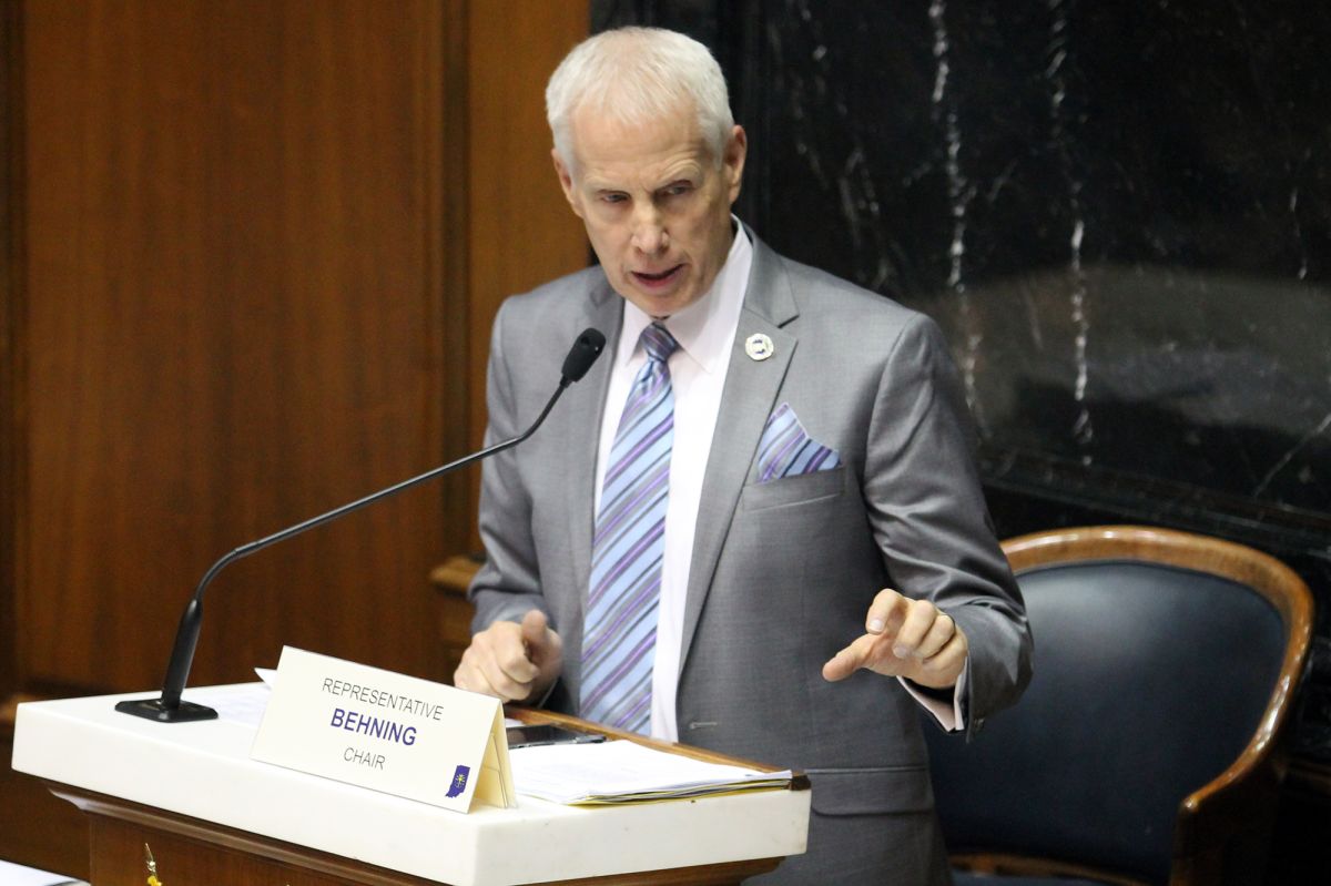 Bob Behning, a White man, is standing behind a podium, lifting his hand and speaking into a microphone.
