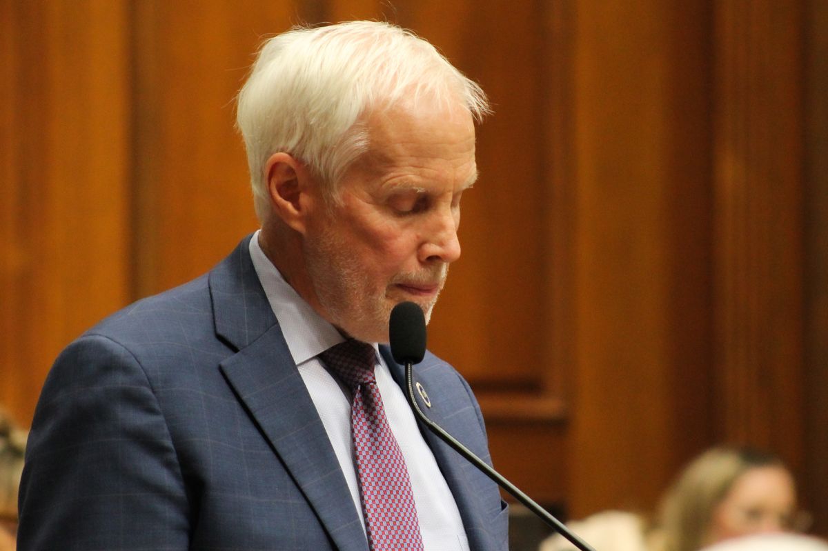Bob Behning wears a suit and has white hair. He looks down while standing in front of a microphone.