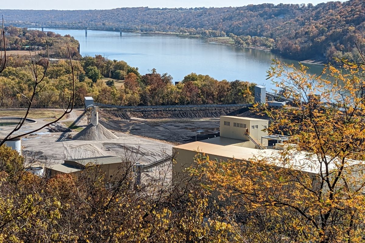 A coal ash pond at the Clifty Creek coal plant in Jefferson County. Bare dirt sits in front of a yellow building. In the background are trees surrounding a body of water with a bridge crossing it.