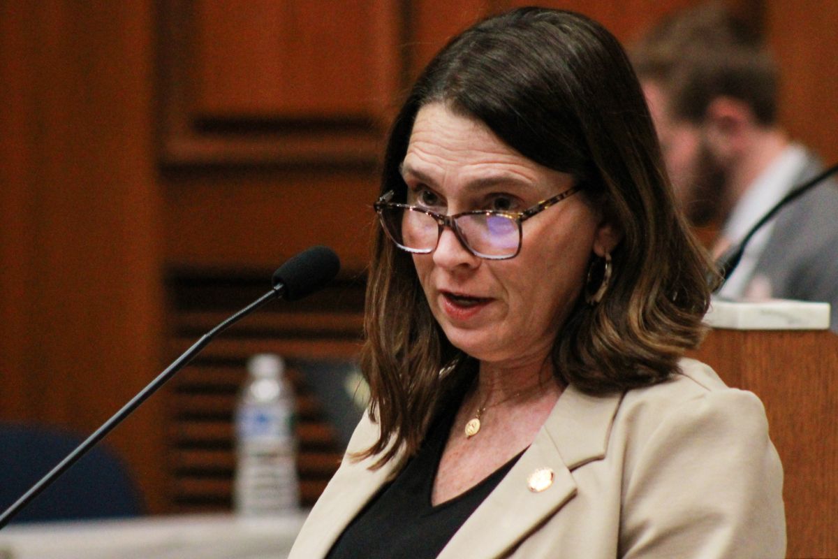 Elizabeth Rowray speaks into a microphone on the House floor. Rowray is a White woman with dark brown hair. She is wearing glasses and a cream colored blazer over a black top.