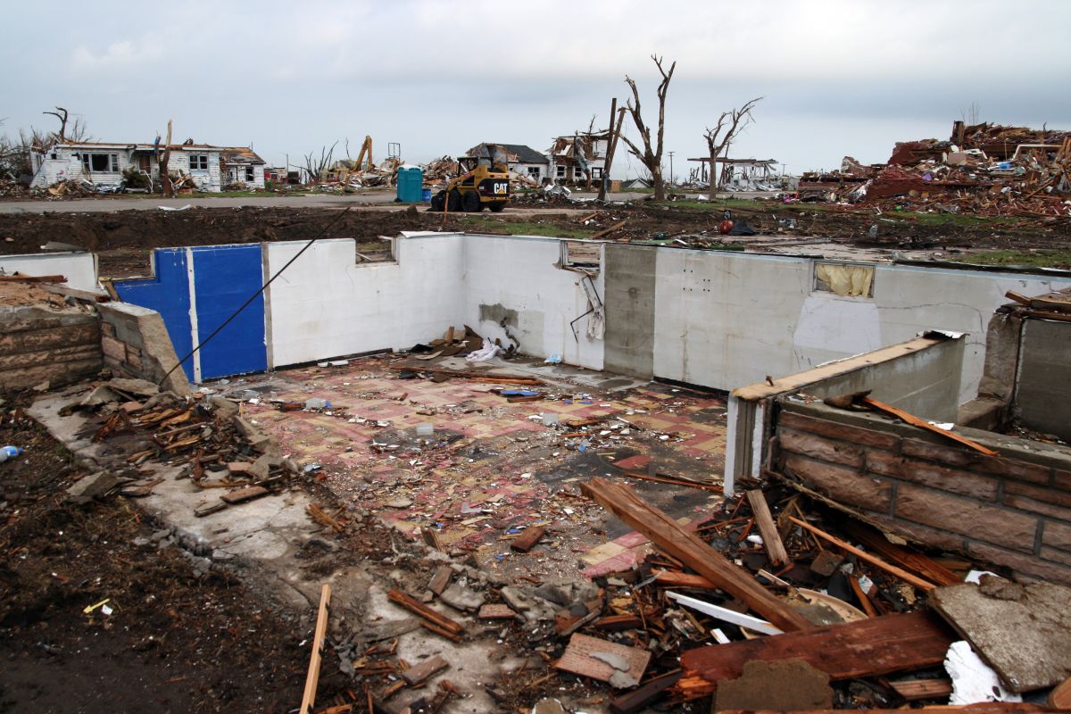 Debris is strewn across a flat landscape from where a home was demolished by a tornado. All that is left of the home is its basement. Across the street, other homes are damaged or partially collapsed. Construction equipment is parked on the street as