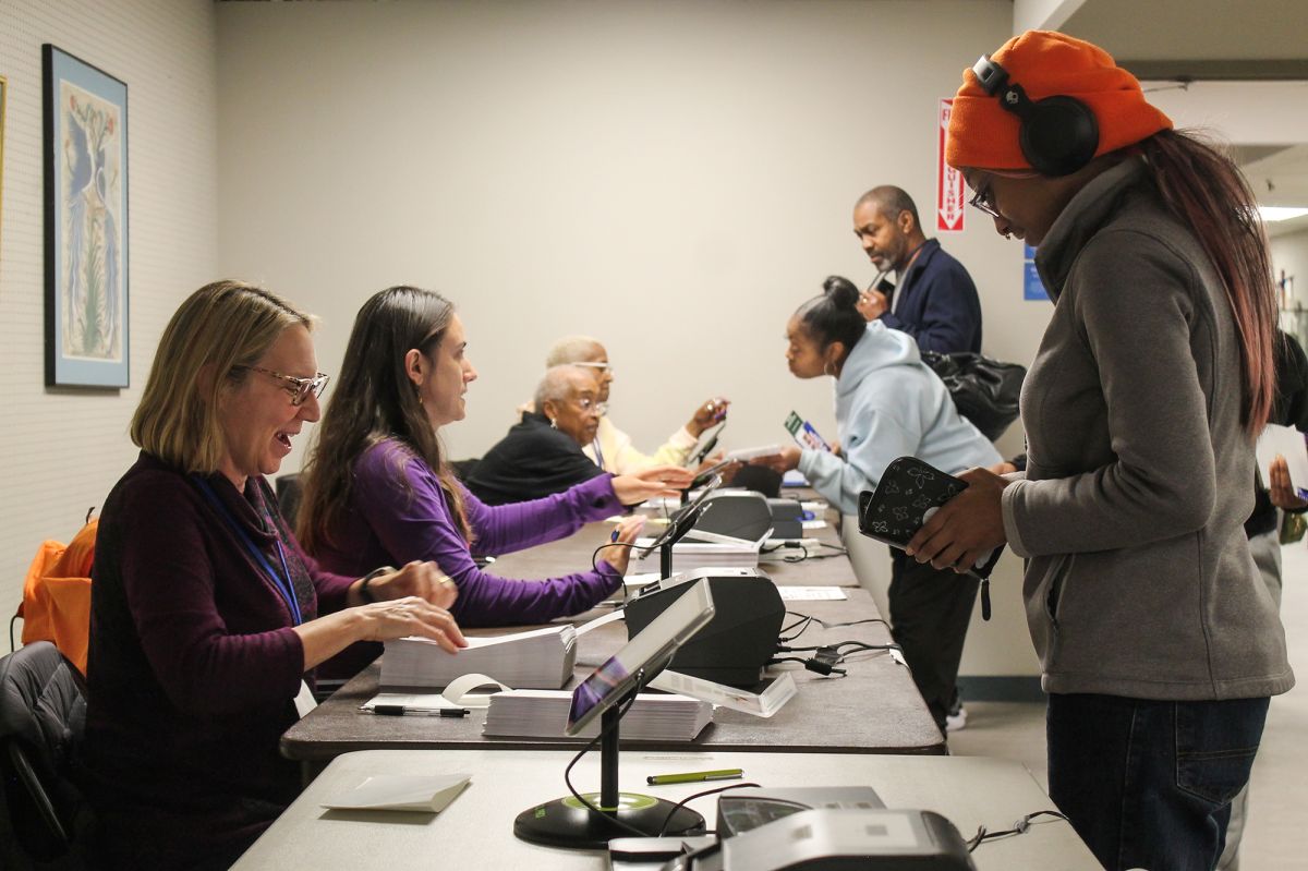 A first-time voter in the 2024 election checks in to vote. She wears an orange hat with headphone on top of it. And she is being checked in by a poll worker, sitting directing across from her working at a tablet.