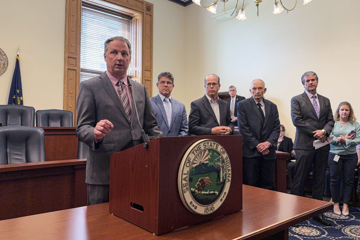 Todd Huston stands a lectern speaking while other Republican leaders stand behind him and off to the side. Huston is a White man with gray hair. He is wearing a gray suit, pink shirt and a gray, purple and pink striped tie.