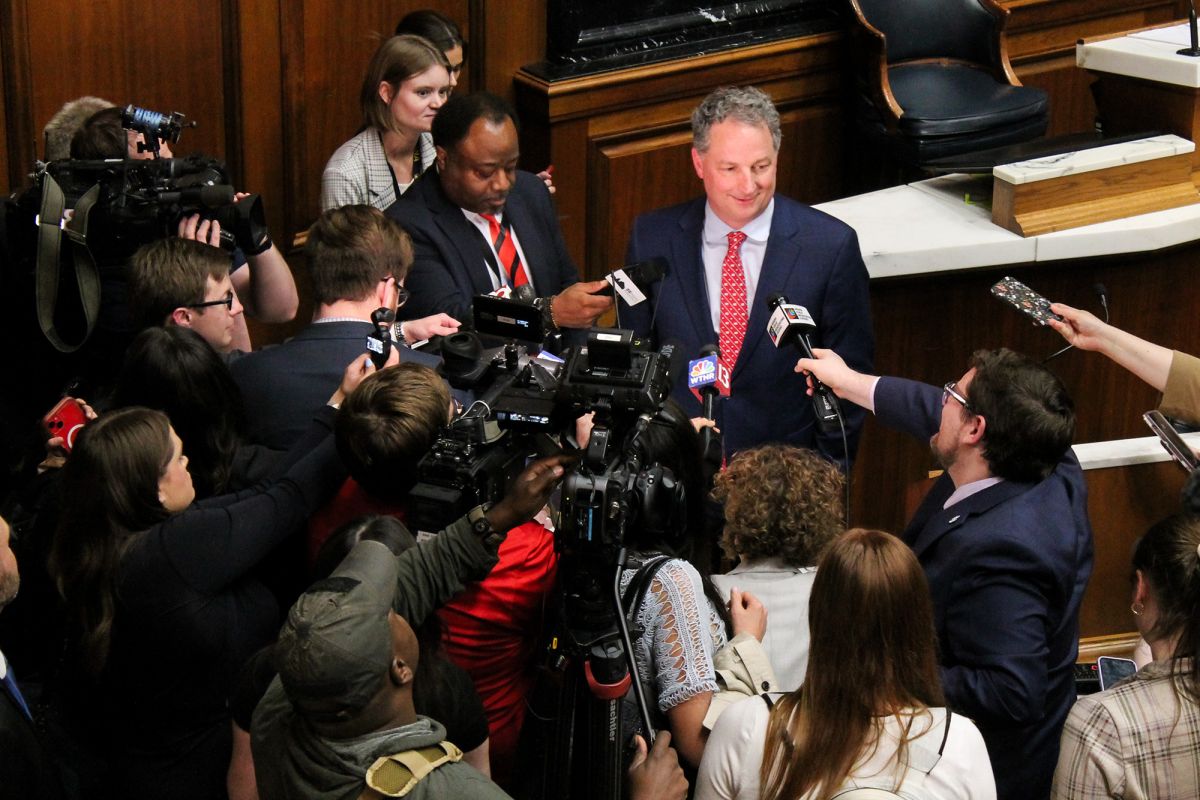 Todd Huston stands in front of a crowd of reporters holding microphones and recorders up at him. Huston is a White man with dark hair. He is wearing a suit and tie.