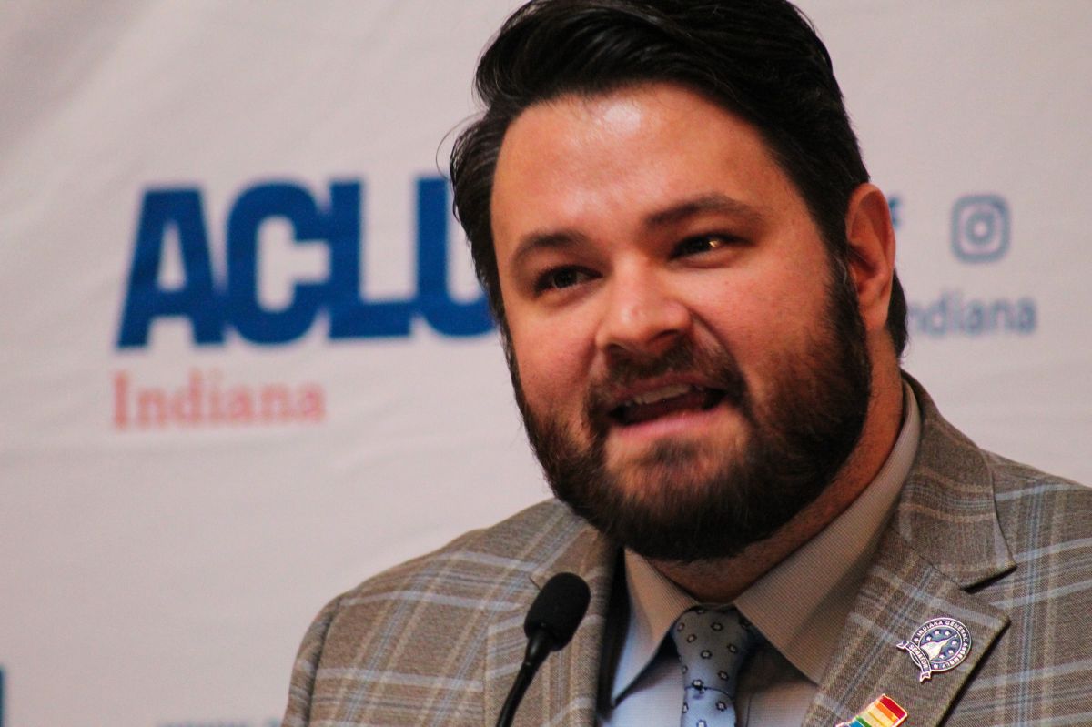 Senator J.D. Ford speaks at an LGBTQ+ Day at the Statehouse. He is a White man with a beard and mustache. He wears a rainbow Indiana pin and plaid suit.
