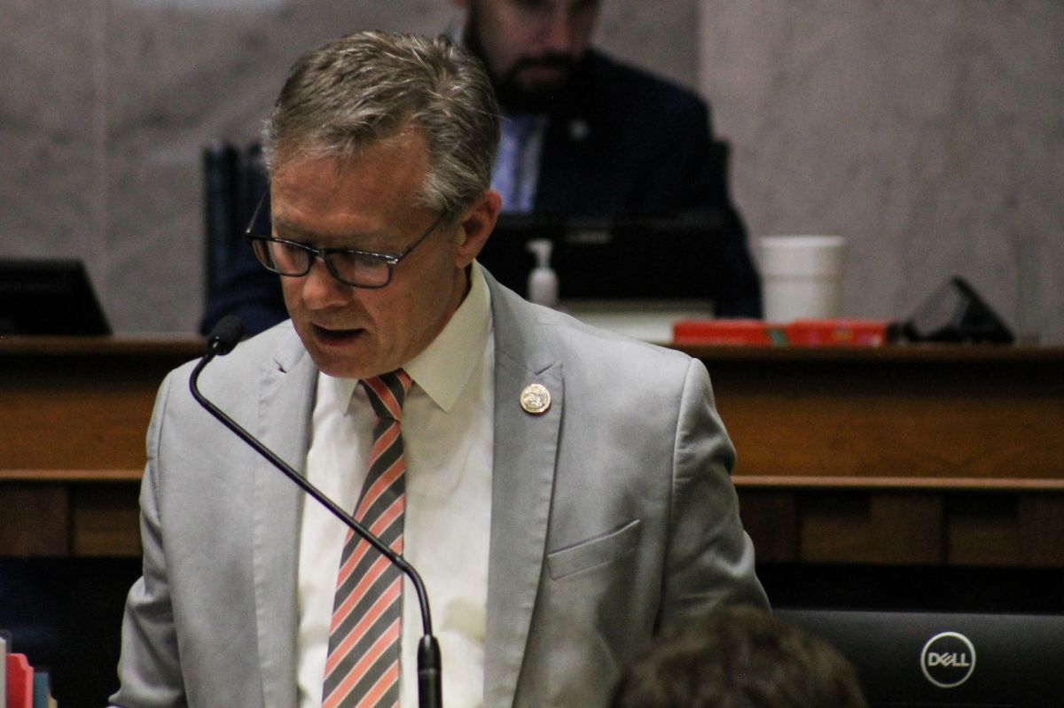 Senator Jeff Raatz wears a salmon and gray striped tie. He stands at a podium and looks down and to the side while speaking into a microphone