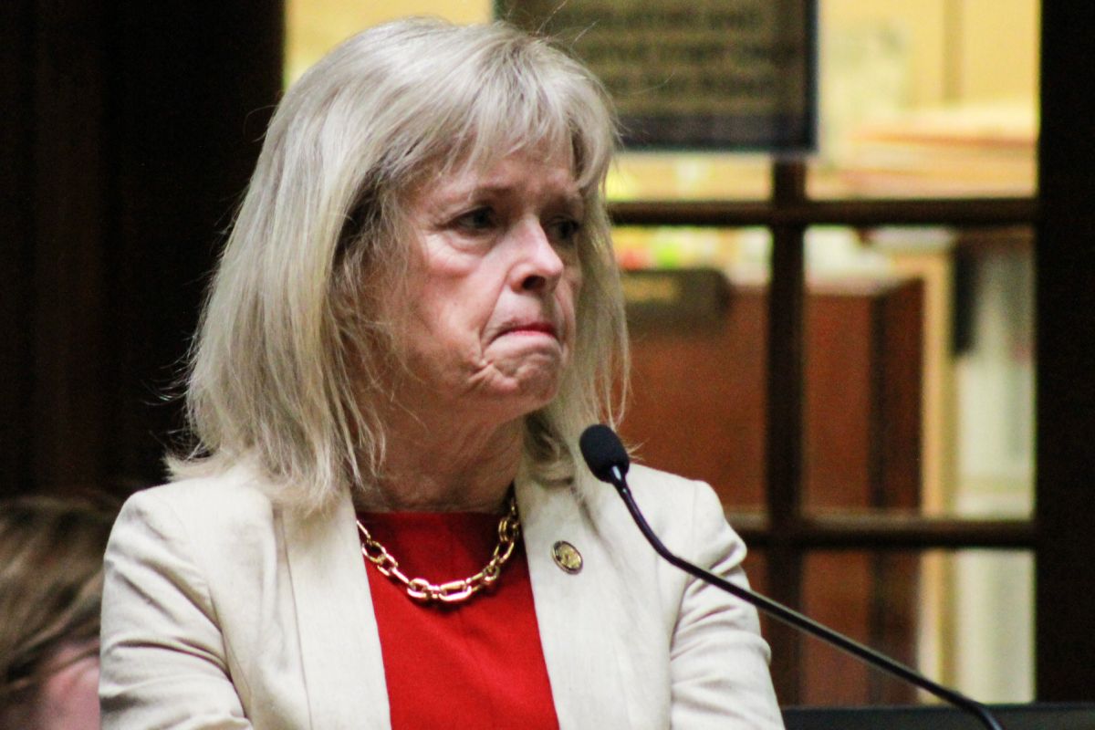 Liz Brown frowns as she speaks on the Senate floor. Brown is a White woman with white and blonde hair. She is wearing a white jacket over a red top.