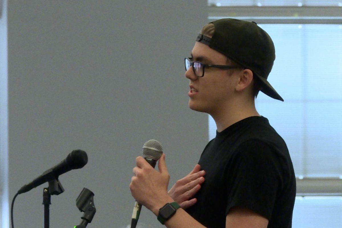 Max Pritchett speaks while holding a microphone. He wears a black t-shirt with a baseball cap and glasses. His right hand is held over his chest.