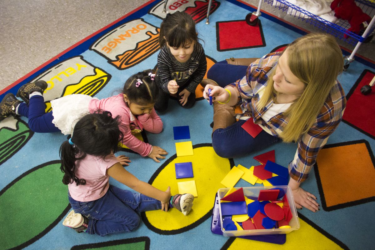 Three young students sort colorful tiles based on their shape while sitting on a mat with various shapes in different colors