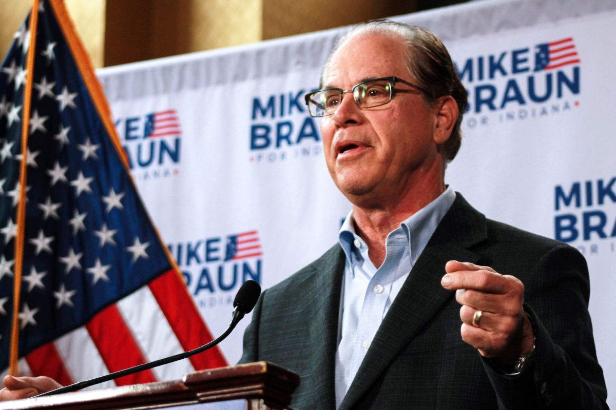 Mike Braun speaks at a lectern on a stage. Behind him are an American flag and a backdrop with his campaign logo. Braun is a White man, balding, with gray hair. He is wearing glasses and a suit.