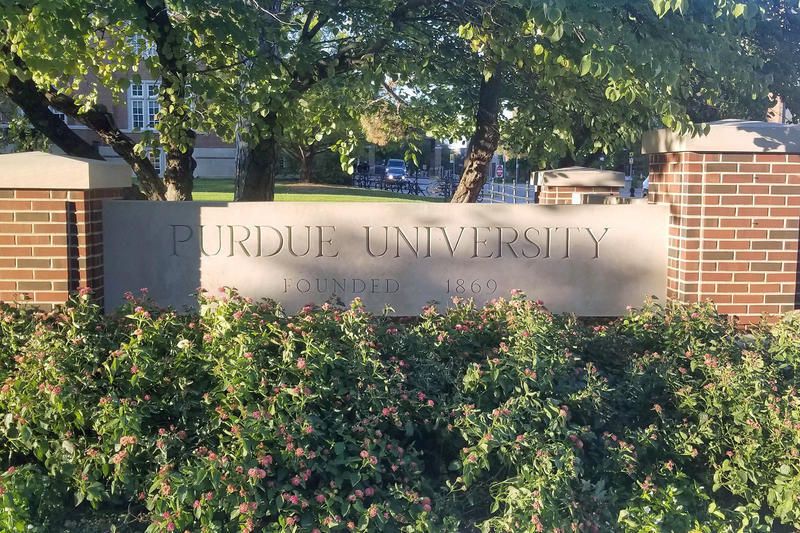 A brick and concrete sign for Purdue University is framed by trees and foliage.