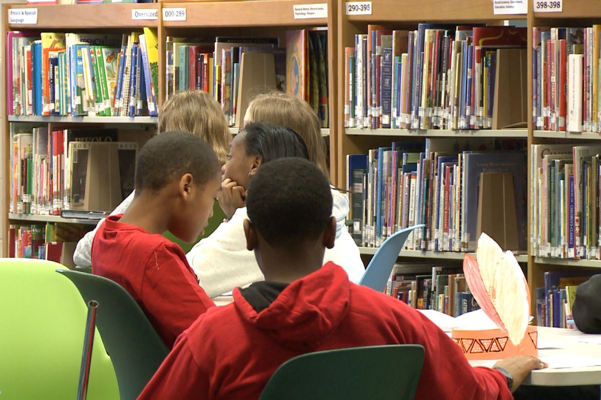 Students sit at ables in a library surrounded by shelves of books.