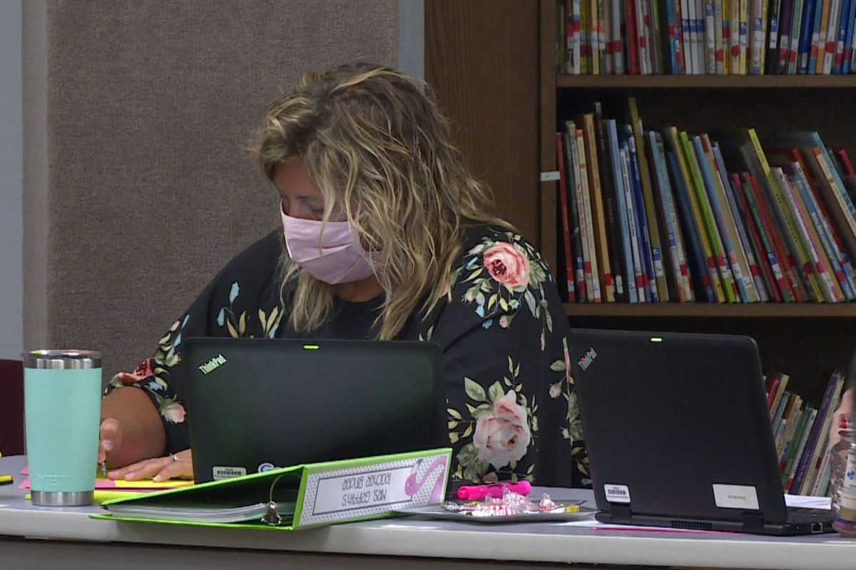 An elementary school teacher wearing a mask sits at a desk and takes notes.