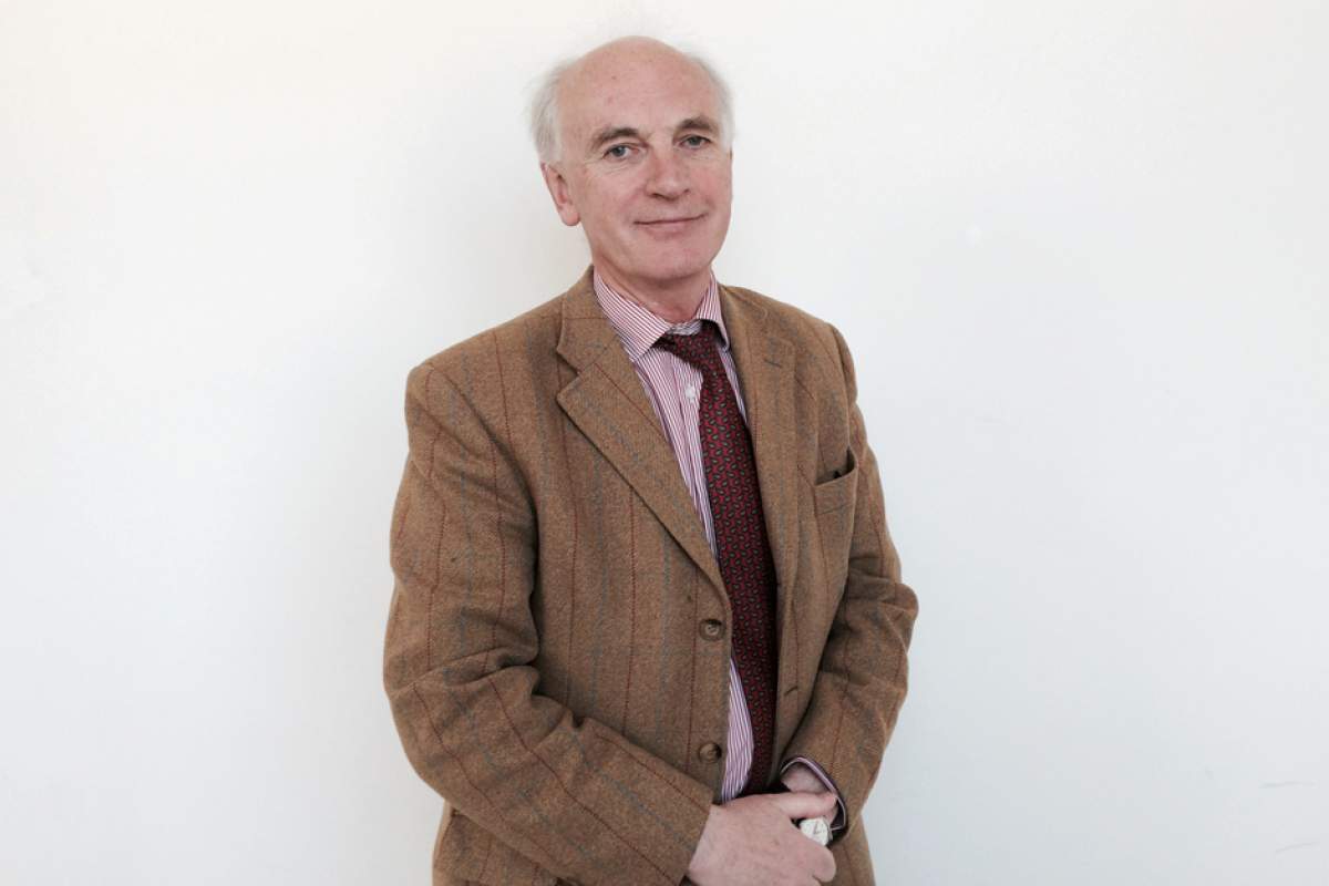 Sir Hew Strachan looking at camera, wearing brown jacket with red stripes, red tie, and dress shirt open at the neck.