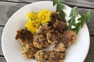 round fried foods on a white plate with yellow dandelion flowers and few jagged leaves