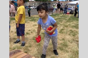 young girl holding red bean bags, about to toss one. browning grass, an apartment complex and other people in the background