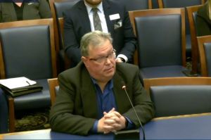 There is a man wearing a grey blazer and white dress shirt while he is standing at a podium. There are people behind him standing beside each other looking at him speak. 