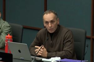 Gary Byrne speaks into a microphone at a lectern in the Senate. Byrne is a White man, mostly bald. He is wearing reading glasses and a dark suit with a blue shirt and a red tie.