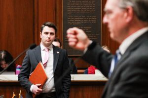 Andrew Ireland stands on the House floor looking at Matt Pierce, who is in the foreground, standing in profile and raising his hand while he speaks. Ireland is a White man with dark hair, wearing a suit and tie. Pierce is a White man with gray hair
