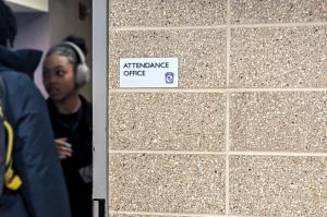A small silver plaque that says "attendance office" is posted on a tan brick wall and students are visible beyond the edge of the doorway