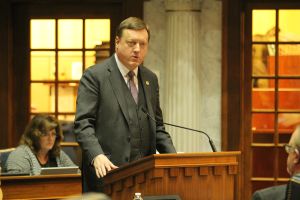 Senator Jon Ford testifies on the floor of the Indiana Senate. He is a White man, bald wearing an navy suit. He stands in front of a microphone.