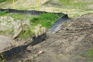 A coal ash pond at the Clifty Creek coal plant in Jefferson County. Bare dirt sits in front of a yellow building. In the background are trees surrounding a body of water with a bridge crossing it.
