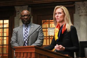 Lindsay Weaver sits in front of a microphone. Weaver is a White woman with dark blonde hair. She is wearing a gray blazer over a black turtleneck.