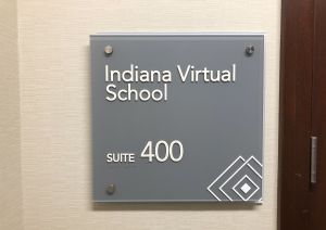 An empty school hallway is lined by lockers. A digital clock at the top of the hallway reads the time and date. The fluorescent lights running down the hallway reflect off the linoleum flooring.